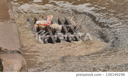 Rainwater runoff carrying urban waste flowing into a clogged storm drain Rainwater runoff carrying urban waste flowing into a clogged storm drain 136140296
