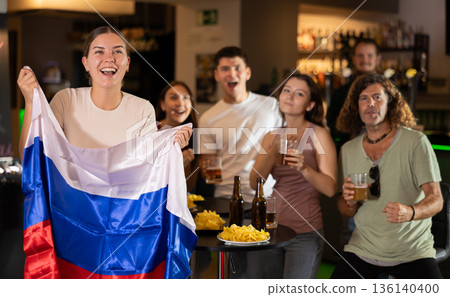 Football fans gesturing emotionally after goal scored by favorite team while watching championship match on TV in sports bar with Russian flag 136140400