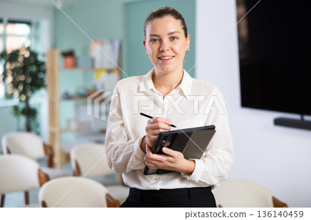 Positive friendly young girl secretary with digital tablet in hands gesture inviting visitors to enter in office Positive friendly young girl secretary with digital tablet in hands gesture inviting visitors to enter in office 136140459
