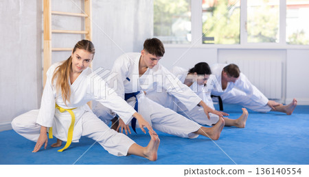 Young woman wearing kimono practicing leg stretching position in sports hall Young woman wearing kimono practicing leg stretching position in sports hall 136140554