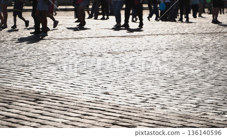Crowd of people walking on cobblestone street creating shadows in bright sunlight 136140596