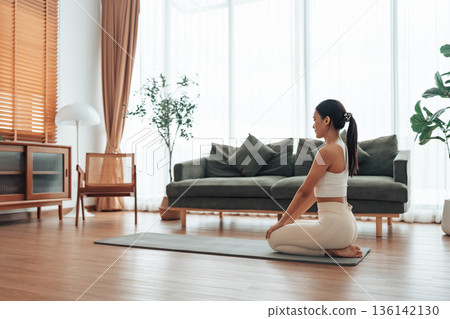 Asian woman sits on an exercise mat, getting ready to exercise in living room at home 136142130