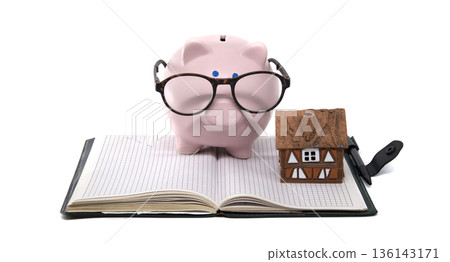 Smart piggy bank with glasses and house model on open notebook, symbolizing financial planning Smart piggy bank with glasses and house model on open notebook, symbolizing financial planning 136143171