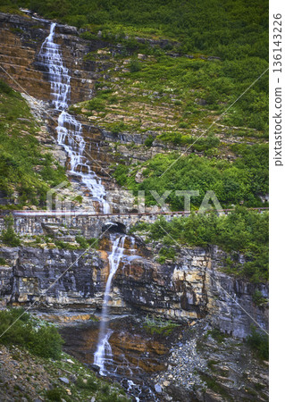 Waterfall Cascading Over Rocky Cliff with Scenic Mountain Road and Lush Greenery Waterfall Cascading Over Rocky Cliff with Scenic Mountain Road and Lush Greenery 136143226
