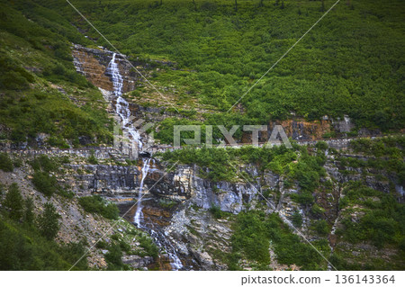 Waterfall Flowing Beside Scenic Mountain Road and Lush Green Hillside 136143364