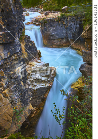 Waterfall Flowing Through Rocky Canyon with Turquoise River and Lush Greenery 136143531