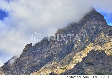 Majestic Rocky Mountain Peak with Dramatic Cloud Cover and Sunlit Cliff Faces 136143581