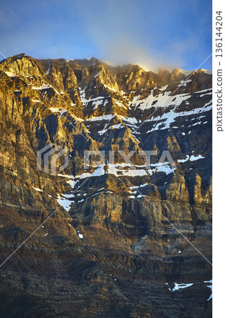 Golden Sunrise Light on Rugged Mountain Cliff with Snow and Clouds 136144204