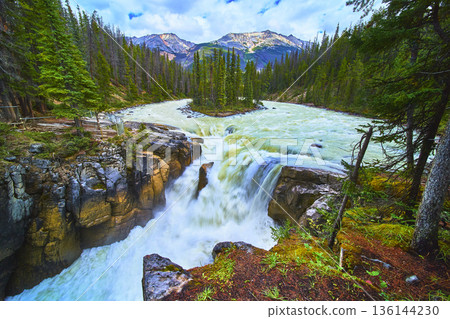 Waterfall Flowing Over Rocky Cliff Surrounded By Pine Trees And Majestic Mountain Peaks Waterfall Flowing Over Rocky Cliff Surrounded By Pine Trees And Majestic Mountain Peaks 136144230