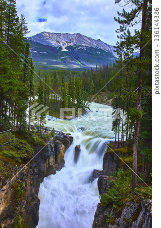 Waterfall And River Flowing Through Forest With Mountain In Background 136144386