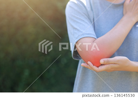 Close-up of woman hand holding painful injured elbow with red inflamed spot. Concept of joint bone pain, muscle injury, arthritis, tendonitis, or ache from sports or work in outdoor background. Close-up of woman hand holding painful injured elbow with red inflamed spot. Concept of joint bone pain, muscle injury, arthritis, tendonitis, or ache from sports or work in outdoor background. 136145530