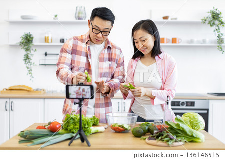 A happy couple prepares a meal in their kitchen, filming a cooking tutorial for their online audience A happy couple prepares a meal in their kitchen, filming a cooking tutorial for their online audience 136146515