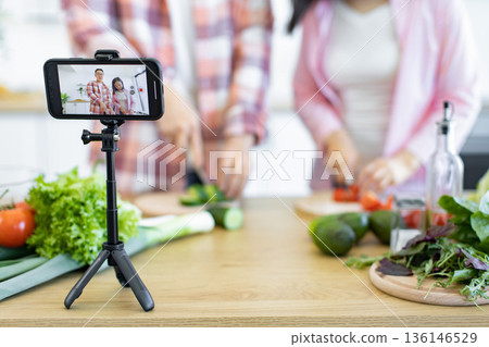 A couple films a cooking vlog in their kitchen, using a smartphone on a tripod to record the process 136146529