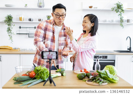 An Asian couple is filming a cooking video in their bright, modern kitchen, preparing fresh vegetables for a healthy meal An Asian couple is filming a cooking video in their bright, modern kitchen, preparing fresh vegetables for a healthy meal 136146532