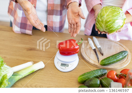 A red bell pepper is weighed on a kitchen scale with other fresh vegetables nearby 136146597