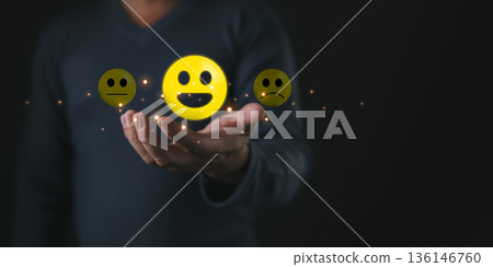 A person holds glowing emoticons showing happy,neutral,and sad faces symbolizing think positive mindset,customer satisfaction,feedback,emotions,evaluation,and choice in expressing experiences. 136146760