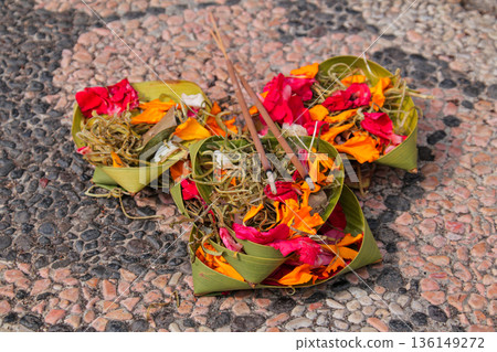 Balinese Canang Sari daily offerings, three baskets, flower petals, and incense sticks placed on a rough stone ground. Balinese Canang Sari daily offerings, three baskets, flower petals, and incense sticks placed on a rough stone ground. 136149272