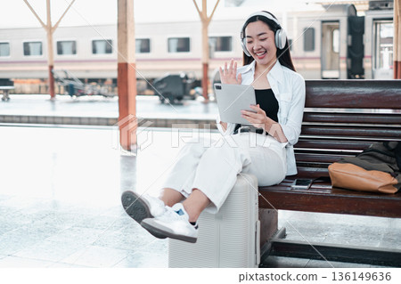 Young woman with headphones using a tablet at a train station, sitting on luggage, smiling and waving. 136149636