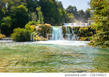 Waterfalls at Skradinski Buk, national nature park Krka, Croatia. Flowing water in beautiful nature, green plants and trees. Sunny summer day. Waterfalls at Skradinski Buk, national nature park Krka, Croatia. Flowing water in beautiful nature, green plants and trees. Sunny summer day. 136149823