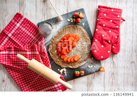 Homemade galette pie cake with strawberries and almond, placed on black desk and wooden table. Red towel with cooking tools on the side Homemade galette pie cake with strawberries and almond, placed on black desk and wooden table. Red towel with cooking tools on the side 136149941