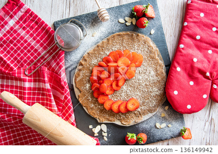 Homemade galette pie cake with strawberries and almond, placed on black desk and wooden table. Red towel with cooking tools on the side 136149942