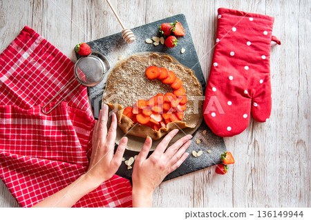 Galette with strawberries preparation, detail of hands of cooker. Pie cake placed on black desk and wooden table with cooking tools on the side. Galette with strawberries preparation, detail of hands of cooker. Pie cake placed on black desk and wooden table with cooking tools on the side. 136149944