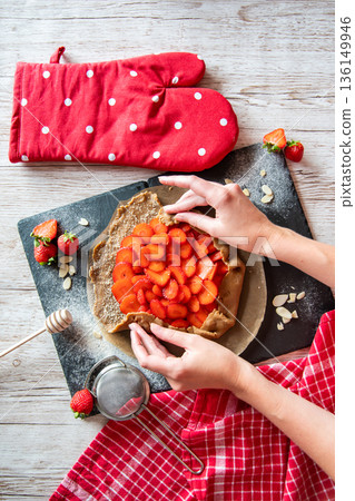 Galette with strawberries preparation, detail of hands of cooker. Pie cake placed on black desk and wooden table with cooking tools on the side. 136149946