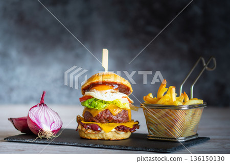 Tasty homemade cheeseburger with french fries placed on black desk, gray background. Cheese, bacon, egg, salad, tomato and onion in light bread. Unhealthy food, concept of fast food. 136150130