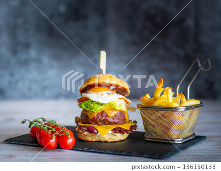 Tasty homemade cheeseburger with french fries placed on black desk, gray background. Cheese, bacon, egg, salad, tomato and onion in light bread. Unhealthy food, concept of fast food. Tasty homemade cheeseburger with french fries placed on black desk, gray background. Cheese, bacon, egg, salad, tomato and onion in light bread. Unhealthy food, concept of fast food. 136150133