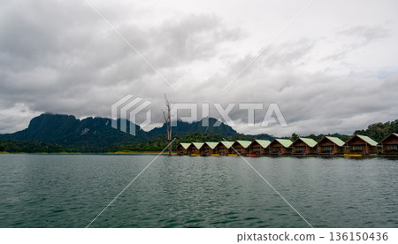 Floating wooden bungalows lined up on the calm waters of Khao Sok Lake in Thailand, surrounded by lush forests and tropical rainforest in a peaceful, remote setting. 136150436