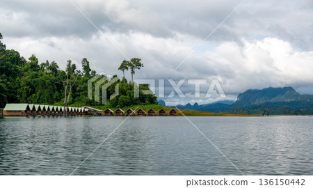 Floating wooden bungalows lined up on the calm waters of Khao Sok Lake in Thailand, surrounded by lush forests and tropical rainforest in a peaceful, remote setting. Floating wooden bungalows lined up on the calm waters of Khao Sok Lake in Thailand, surrounded by lush forests and tropical rainforest in a peaceful, remote setting. 136150442