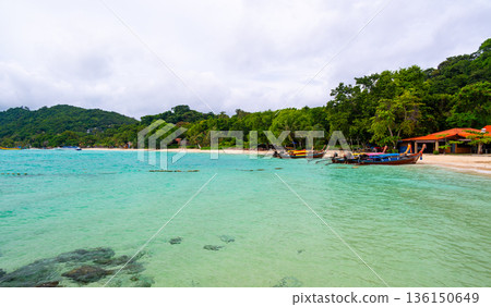 Tropical beach on Ko Phi Phi island in Thailand with palm trees, lush rainforest, and traditional long tail boats anchored along the shore under the bright sun. Tropical beach on Ko Phi Phi island in Thailand with palm trees, lush rainforest, and traditional long tail boats anchored along the shore under the bright sun. 136150649
