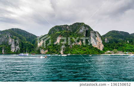 Panoramic wide-angle view of Ko Phi Phi island in Thailand from the sea, surrounded by numerous small and large boats on the clear tropical water under a sunny sky. Panoramic wide-angle view of Ko Phi Phi island in Thailand from the sea, surrounded by numerous small and large boats on the clear tropical water under a sunny sky. 136150652