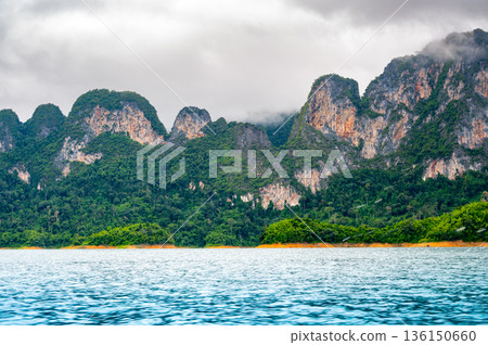 View of Khao Sok Lake in Krabi province, Thailand, with towering limestone cliffs covered in tropical rainforest rising above the calm surface of the lake. View of Khao Sok Lake in Krabi province, Thailand, with towering limestone cliffs covered in tropical rainforest rising above the calm surface of the lake. 136150660