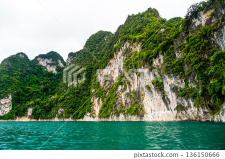 View of Khao Sok Lake in Krabi province, Thailand, with towering limestone cliffs covered in tropical rainforest rising above the calm surface of the lake. 136150666