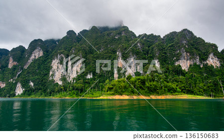 View of Khao Sok Lake in Krabi province, Thailand, with towering limestone cliffs covered in tropical rainforest rising above the calm surface of the lake. 136150683