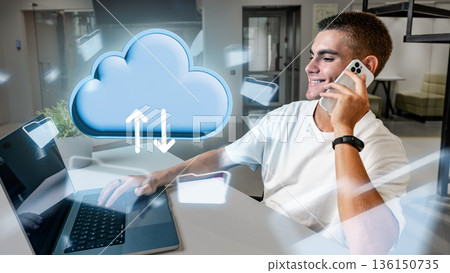Young man uses smartphone while working on laptop in a modern office space during the day Young man uses smartphone while working on laptop in a modern office space during the day 136150735