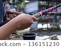Close up of person's hand holding colorful fishing rod at outdoor pond for fishing hobby concept Close up of person's hand holding colorful fishing rod at outdoor pond for fishing hobby concept 136151230