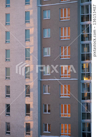 Apartment building with numerous windows on its facade showing sunset light inside. 136153487
