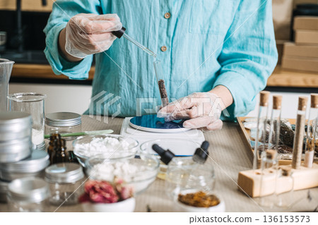 Gloved hands use glass dropper to add liquid to botanical ingredients in test tube on digital scale. Liquid boosters, floral droplets, herbal vials, custom blends. Gloved hands use glass dropper to add liquid to botanical ingredients in test tube on digital scale. Liquid boosters, floral droplets, herbal vials, custom blends. 136153573