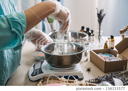 Person sprinkles white powder into metal bowl on digital scale for cosmetic preparation. Floral bath salt layers, apothecary bath mixology, botanical soak formulation, aesthetic dry ingredients mix. 136153577