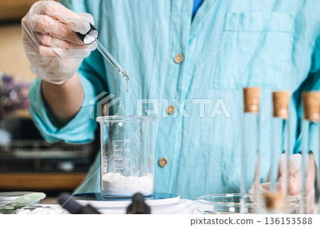 Person uses glass pipette to dispense liquid into beaker containing white powder on digital scale. sustainable beauty lab, zero waste skincare diy, eco-friendly cosmetic packaging,  136153588