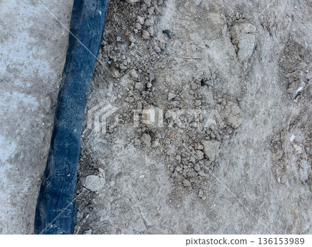 A black erosion control fabric strip is secured over compacted dirt at a construction site. The dry soil surface contains visible rocks and loose particles. The photo highlights the material used to 136153989