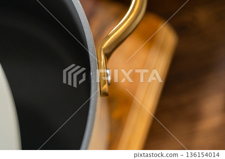 A close up view shows a modern saucepan with a large gold handle resting on a stovetop. The dark interior contrasts with the metallic details. The scene highlights kitchen equipment and cooking tools. 136154014