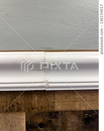 Close view of white baseboard trim running along a painted wall above a wood floor. Soft lighting reveals texture and subtle imperfections in the trim surface. 136154017
