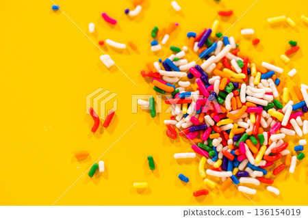Macro shot of multicolored candy sprinkles scattered on a vibrant yellow surface. Perfect for backgrounds, dessert-themed designs, or celebration visuals. 136154019