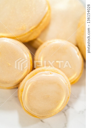 A collection of round sugar cookies decorated with smooth white royal icing and a shimmering gold dust finish. One cookie is being picked up, showing the delicate texture and elegant detailing. These A collection of round sugar cookies decorated with smooth white royal icing and a shimmering gold dust finish. One cookie is being picked up, showing the delicate texture and elegant detailing. These 136154026