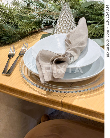 Close-up of a modern dining table with stacked white plates, a beige linen napkin secured with a metallic ring, and soft natural lighting. The setting reflects a clean and elegant design style. Close-up of a modern dining table with stacked white plates, a beige linen napkin secured with a metallic ring, and soft natural lighting. The setting reflects a clean and elegant design style. 136154034