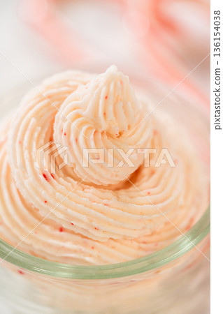 A close-up view highlights the piped swirl of peppermint frosting inside a glass bowl. Red peppermint flecks are visible throughout the pale frosting. The image is softly lit. 136154038