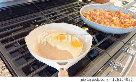 A ceramic pan holds two eggs frying on a stove, with a second skillet of cooked hash in the background. The setup shows a busy breakfast preparation. A ceramic pan holds two eggs frying on a stove, with a second skillet of cooked hash in the background. The setup shows a busy breakfast preparation. 136154055